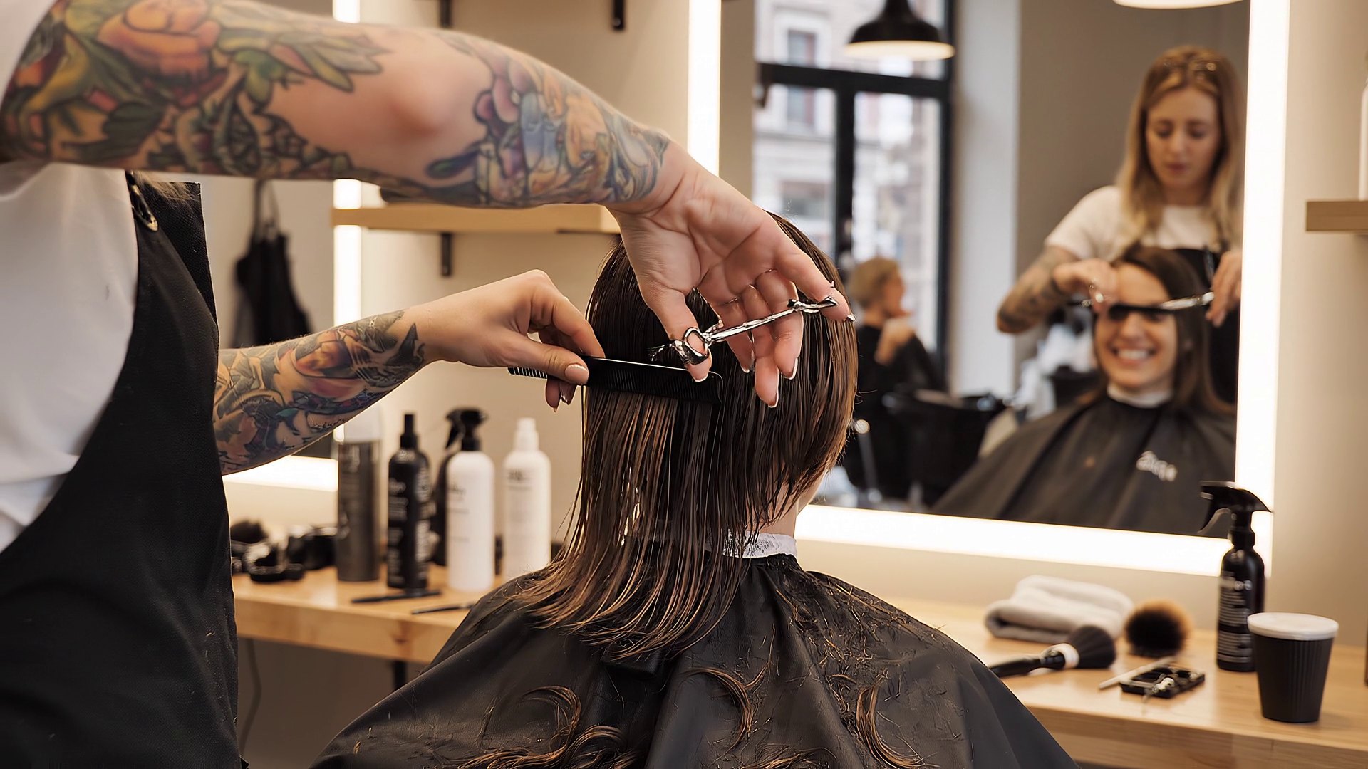 Dans un salon, une coiffeuse peigne et coupe les cheveux d’une cliente qui sourit dans le miroir.